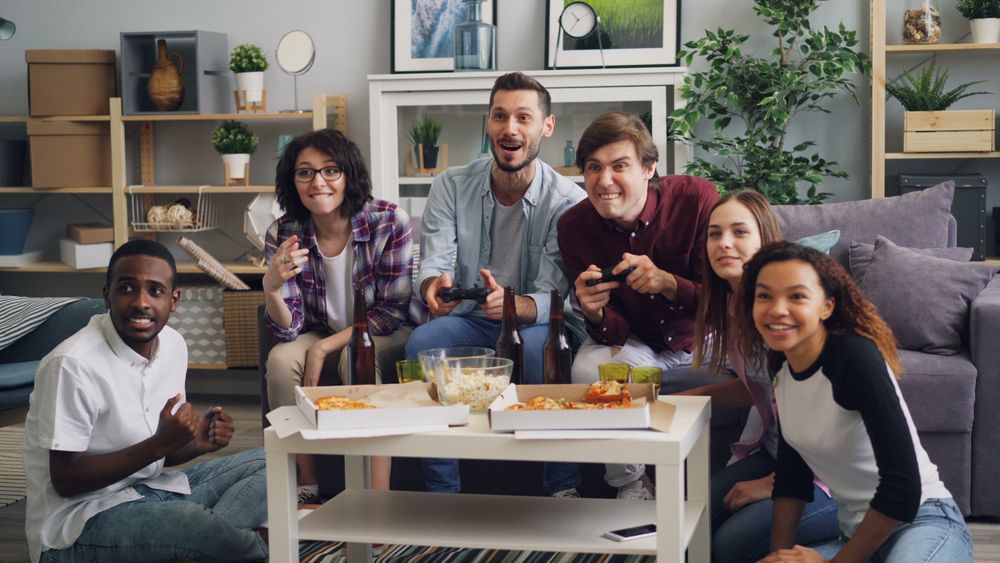 A diverse group of friends having fun playing video games indoors, surrounded by pizza and drinks.